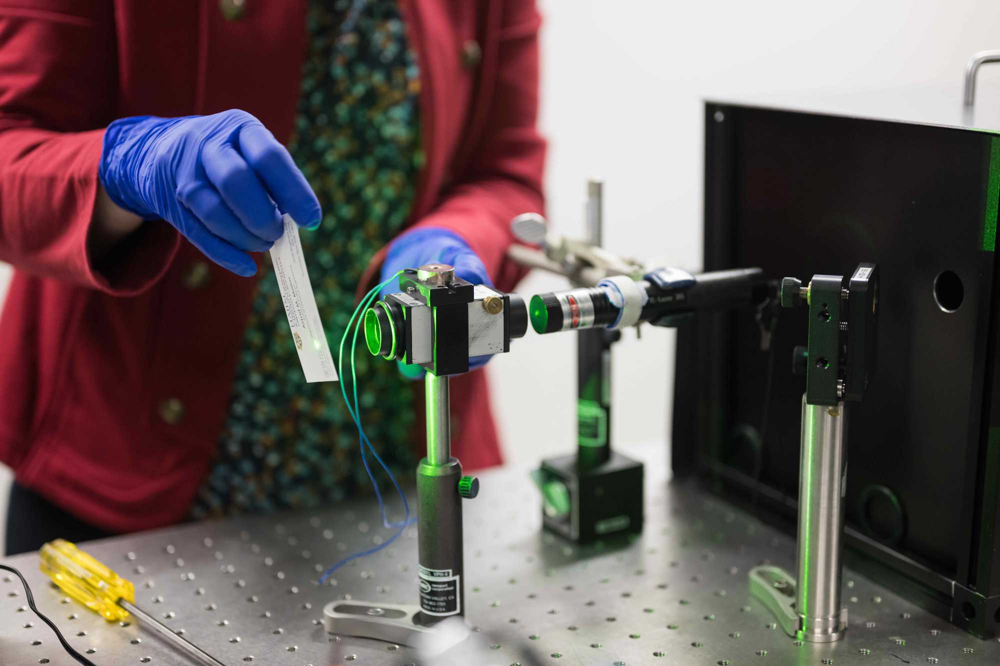 A scientist's gloved hands holds a material in front of a set-up of pulsed lasers to create catalysts.