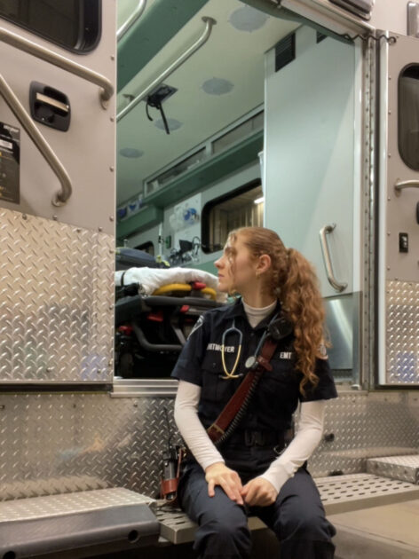 Rachel Witmoyer in an EMT uniform sitting on the back of an ambulance and peering over her shoulder into the vehicle.