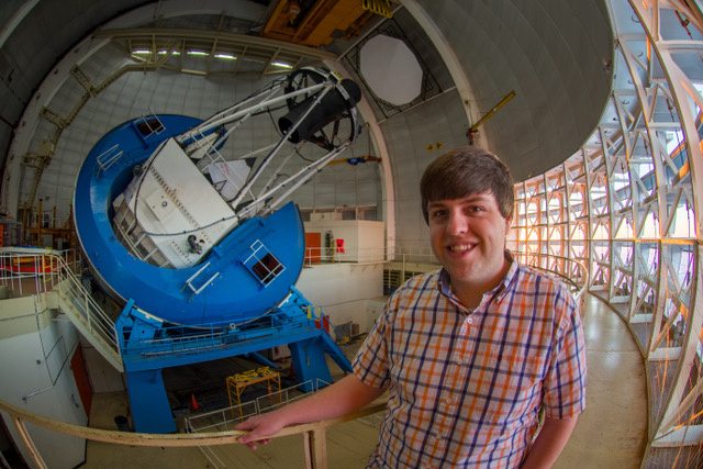 Fish-eye lens view of a Rochester graduate student smiling at the camera with the Dark Energy Spectroscopic Instrument, which is used to detect dark energy, in the background.