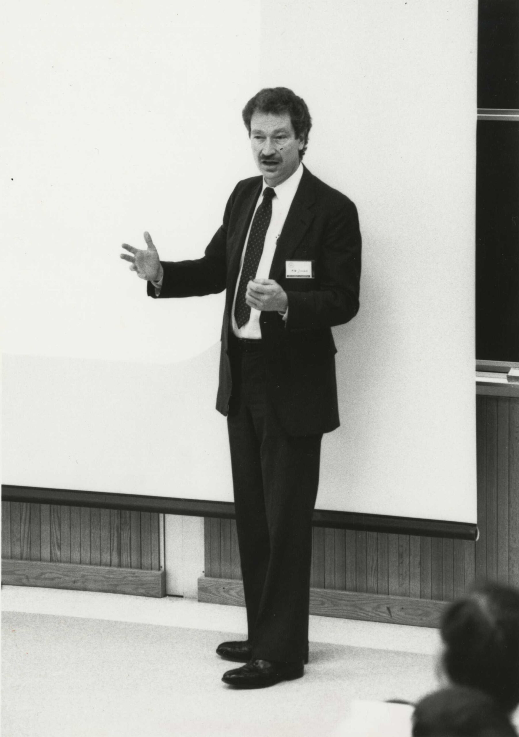 Black and white archival image of Michael Jensen in a suit standing in front of a blank lowered retractable projector screen.