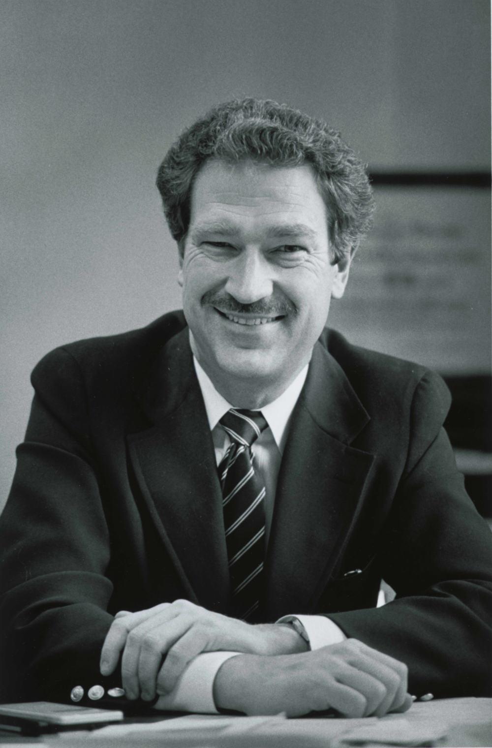 Vertical black-and-white photo of Michael Jensen in a suit with his arms folded on the table in front of him looking at the camera and smiling.