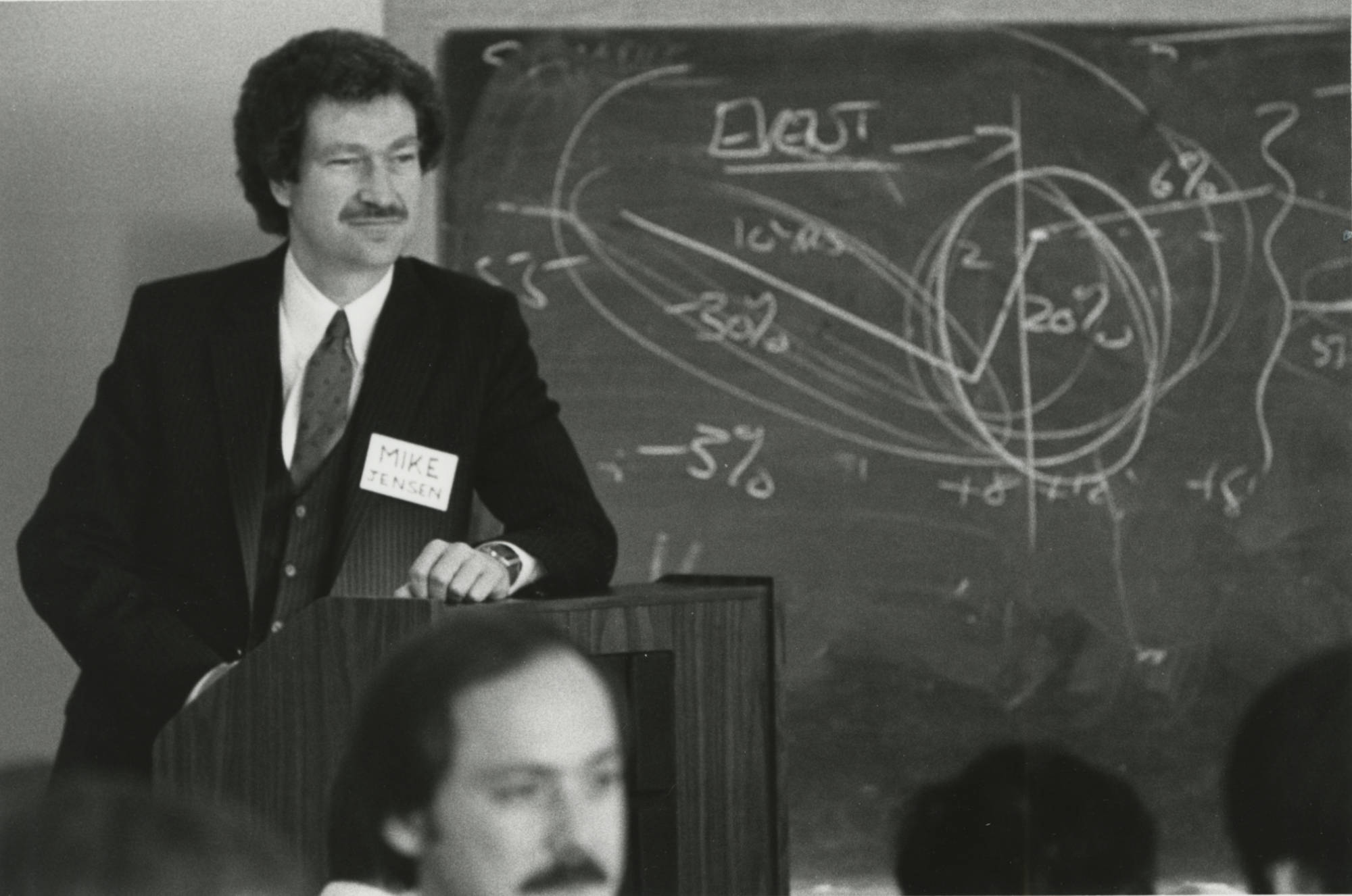 Black and white archival photo of Michael Jensen in a suit and with a big name tag standing at a podium in front of a classroom with a blackboard scribbled with percentages and other figures in the background.
