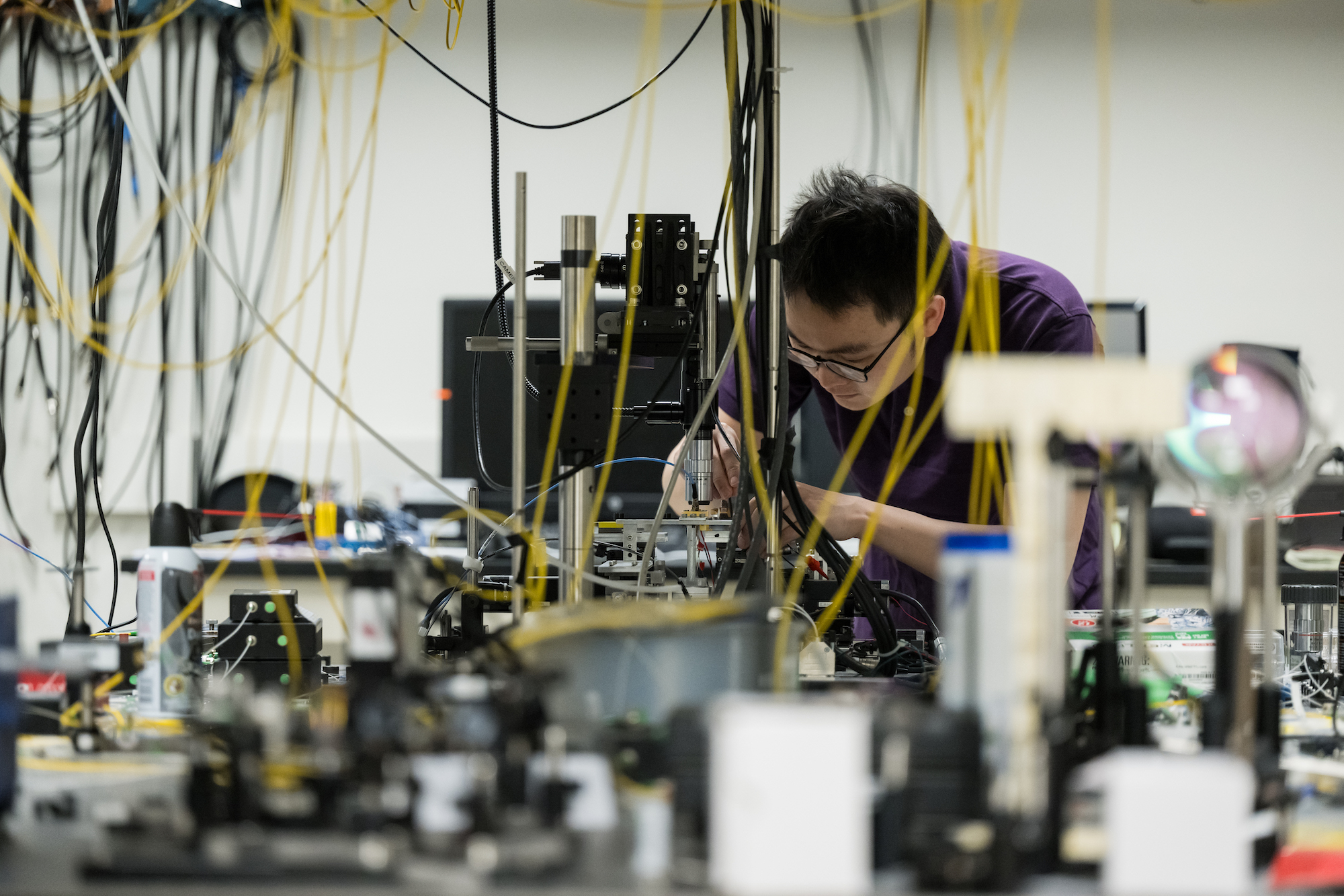 PhD student in an engineering lab, working with a device, surrounded by equipment
