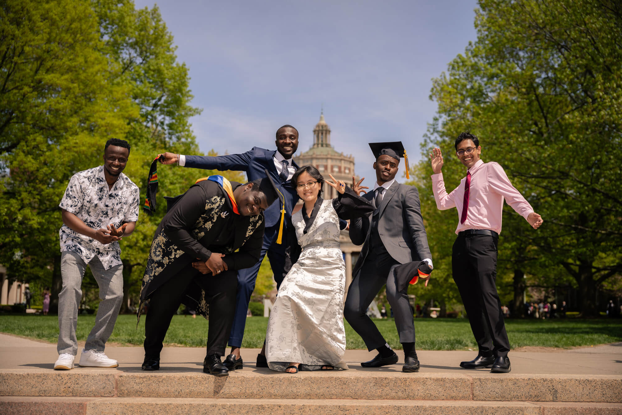 A group of six diverse graduates, dressed in caps, gowns, and formal wear, joyfully pose for a celebratory photo on a sunny day in front of a historic building surrounded by lush green trees. They are smiling and striking playful poses, exuding excitement and pride.