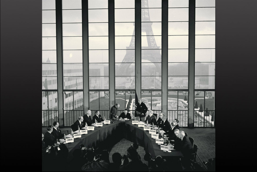 Archival black-and-white image showing the 1956 NATO delegation meeting in a room with Paris's Eiffel Tower visible in the background through the windows.