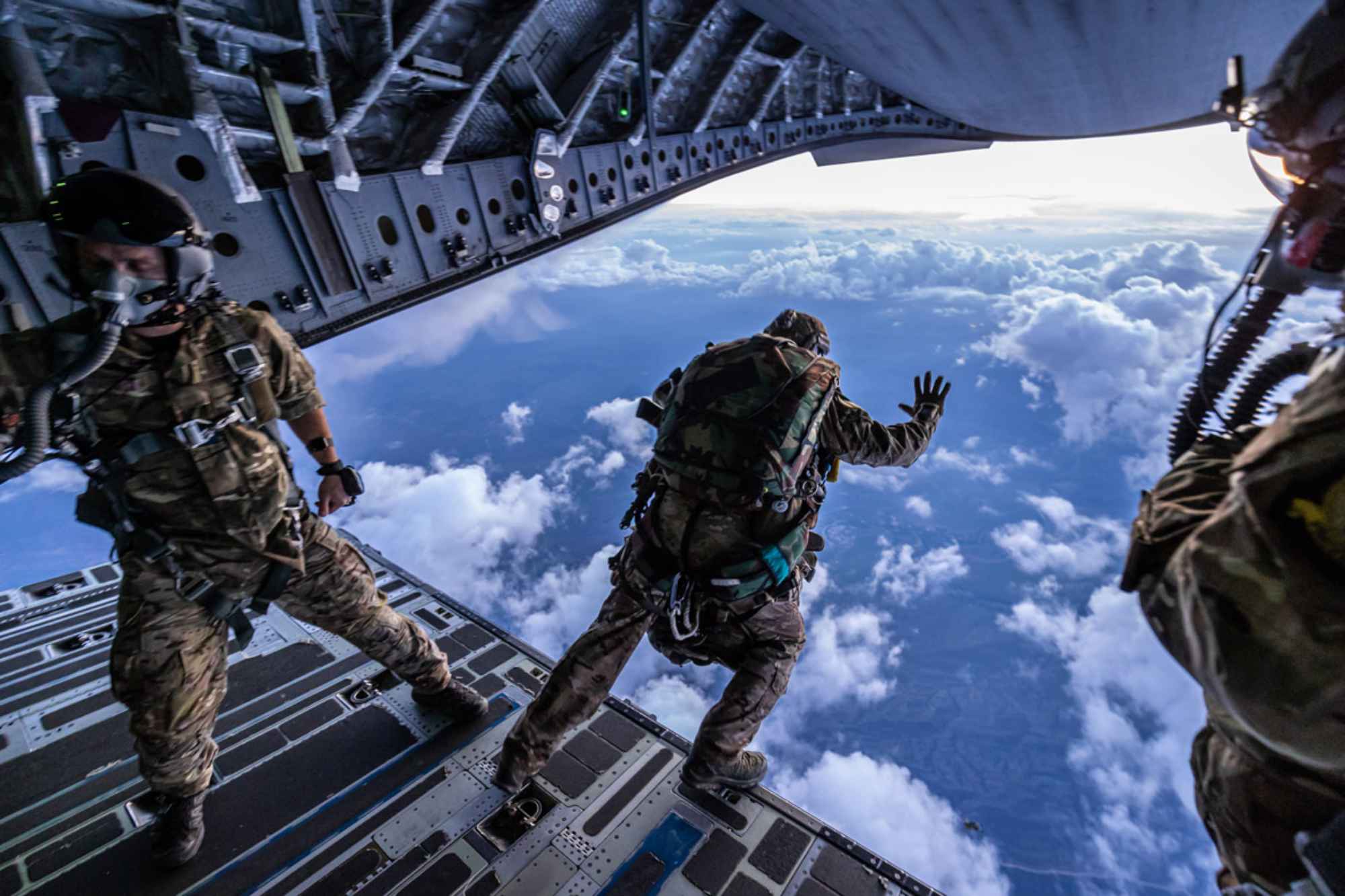 Three British military paratroopers at the back of an aircraft with one leaping into the open sky below.