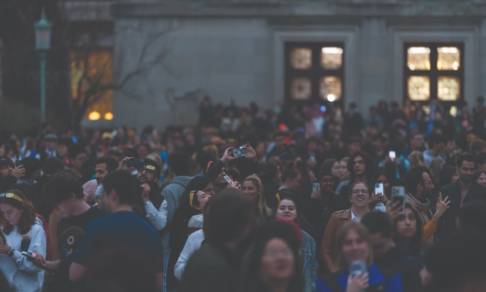 A large crowd of people are gathered outside a building with ornate windows. Some people are raising their phones to take photos or videos. The scene appears to be taking place in the evening or at dusk, with a diverse group of individuals present.