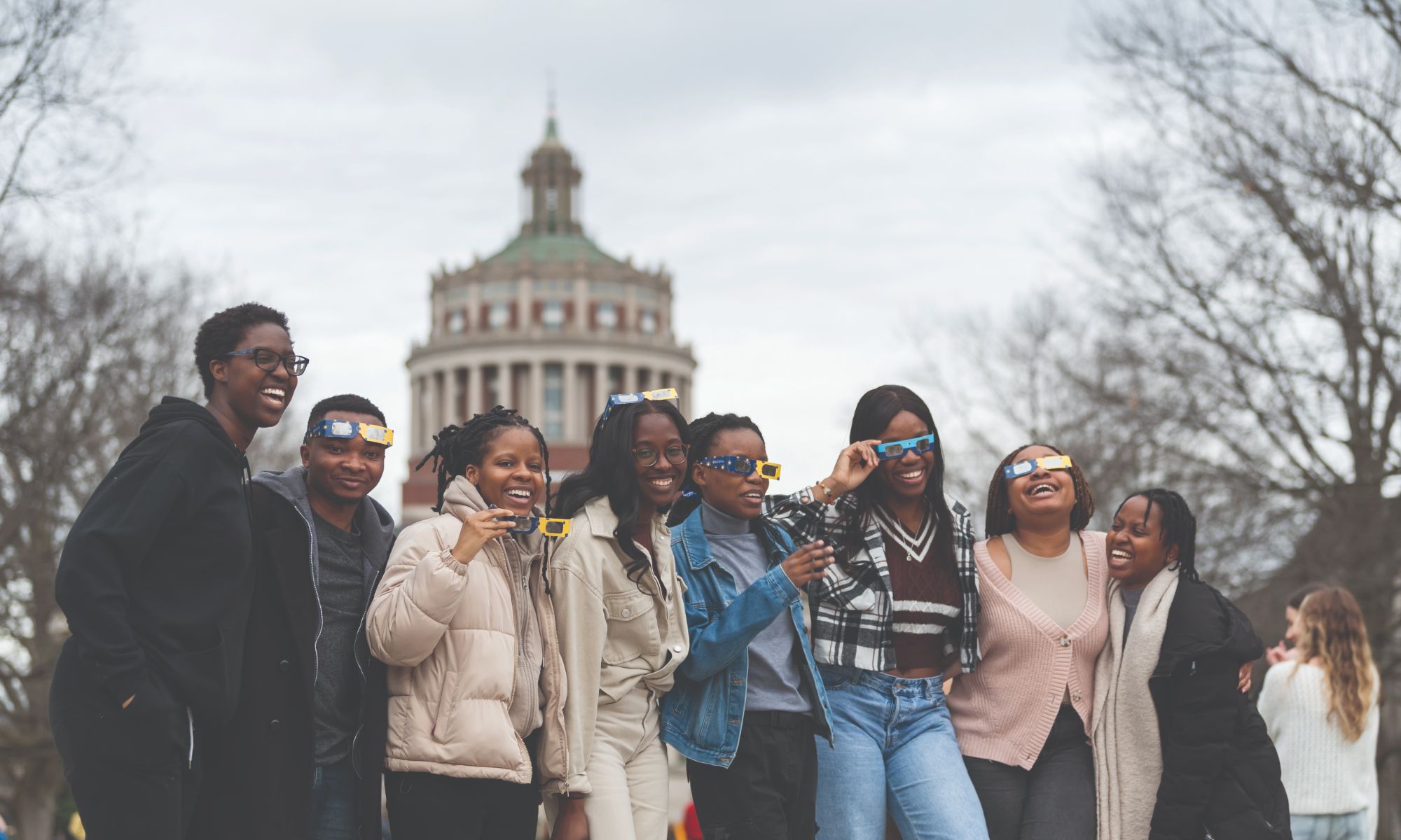 A group of eight people, all smiling and posing for a photo outdoors. Most are wearing sunglasses, some with decorative lenses. A building with a green roof and a dome is visible in the background. They appear happy and are dressed in casual winter clothing.