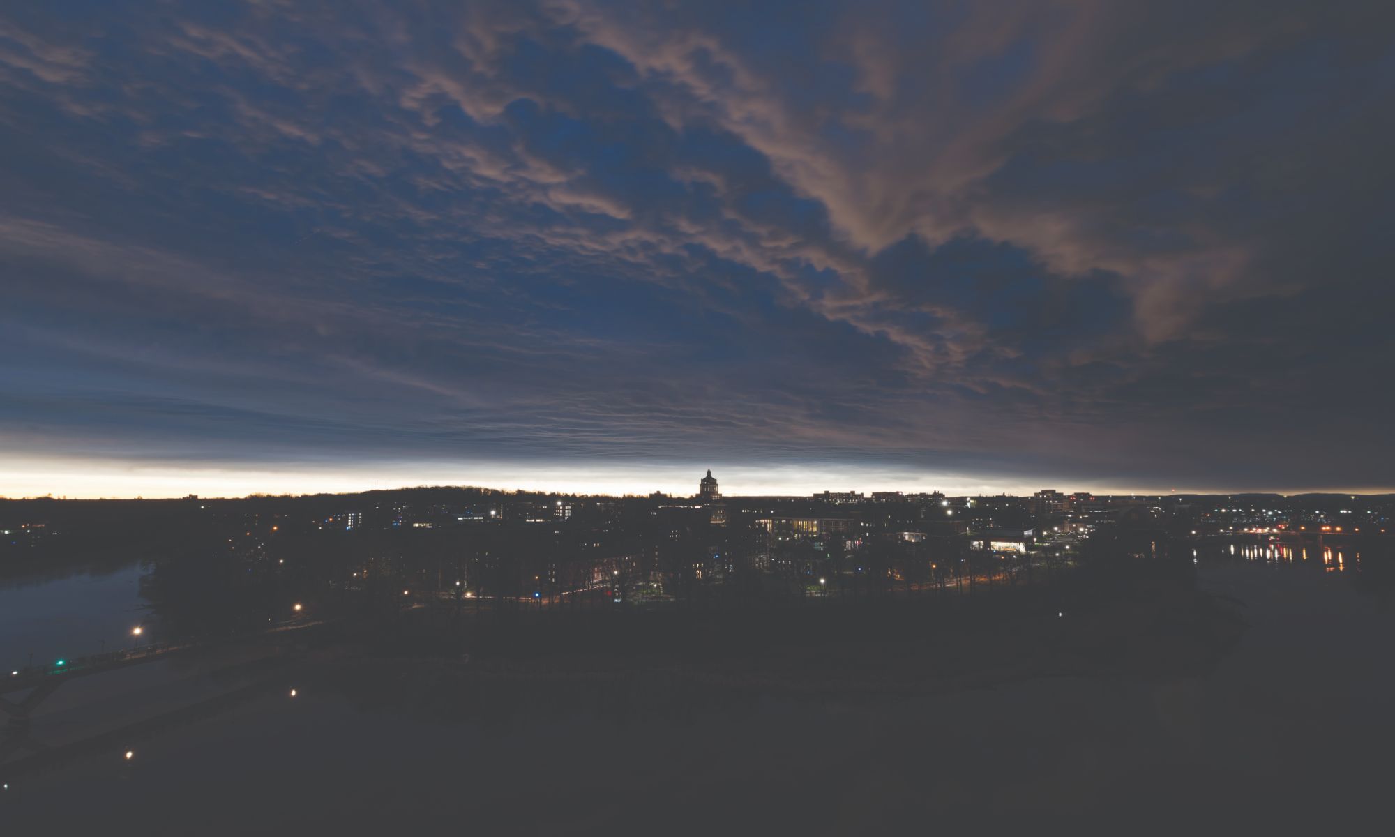 A cityscape at dusk with the skyline illuminated by city lights. The sky is covered with dramatic, dark clouds, with a thin streak of light near the horizon, indicating the setting sun. A river reflects some of the city lights in the foreground.