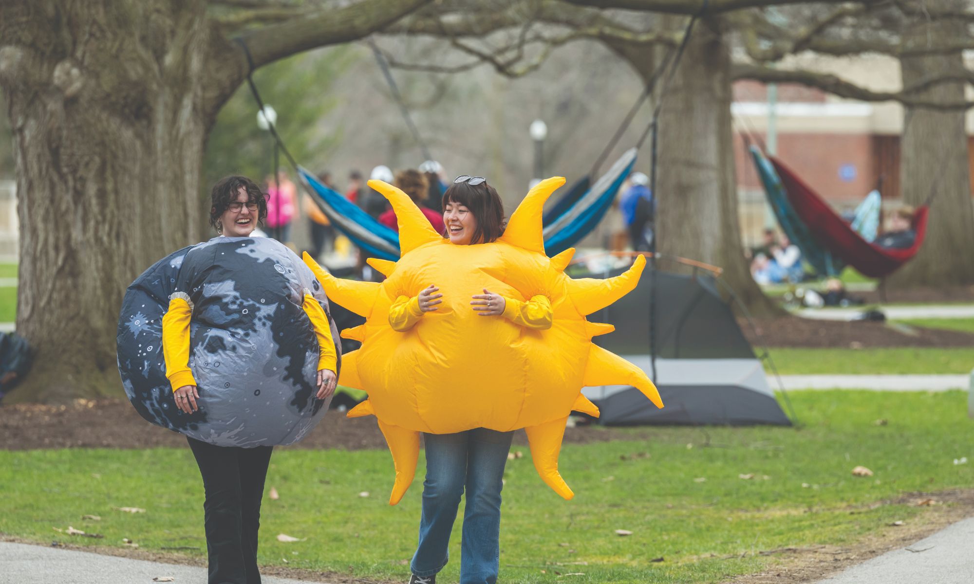 Two people are walking on a lawn, dressed in inflatable costumes; one as a sun and the other as a planet with a cratered surface. They are smiling, and tents and hammocks are set up in the background under large trees. Other people are lounging in the background.