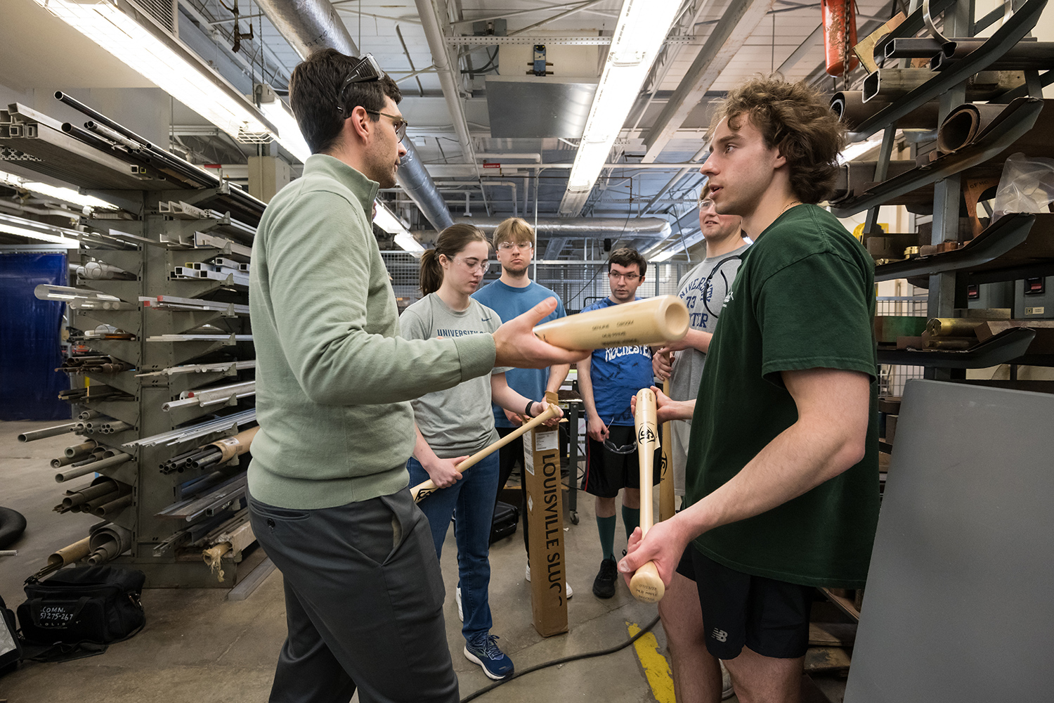 Six people standing in a semi-circle, surrounded by shelving. Three of the people are holding baseball bats and one holds a box labeled LOUISVILL SLUGGER.