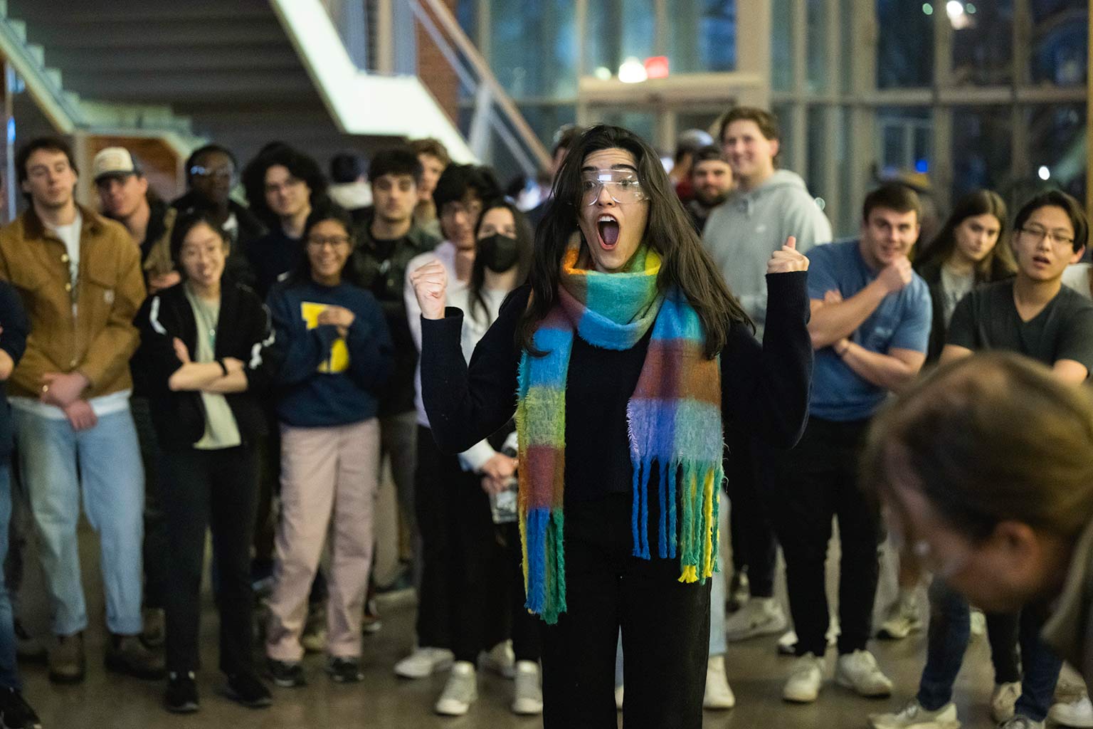 student wearing protective goggles raises her fists in celebration and gives an expression of surprise as she stands in the Goergen Hall atrium surrounded by a crowd of students.