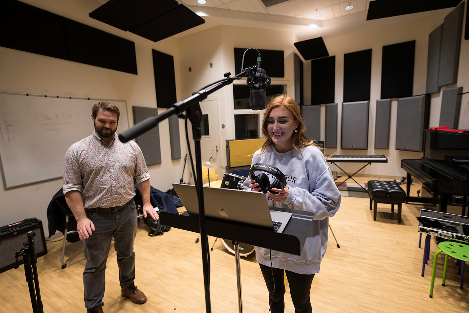 A student stands behind a boom mic and holds a pair of headphones and laughs while looking at a laptop propped onto a music stand while her instructor stands to the side.