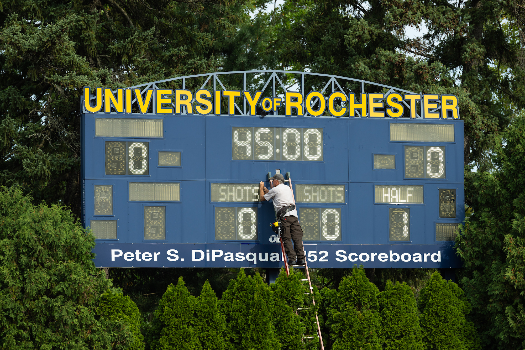 A man atop a ladder fixes a broken panel on a scoreboard 