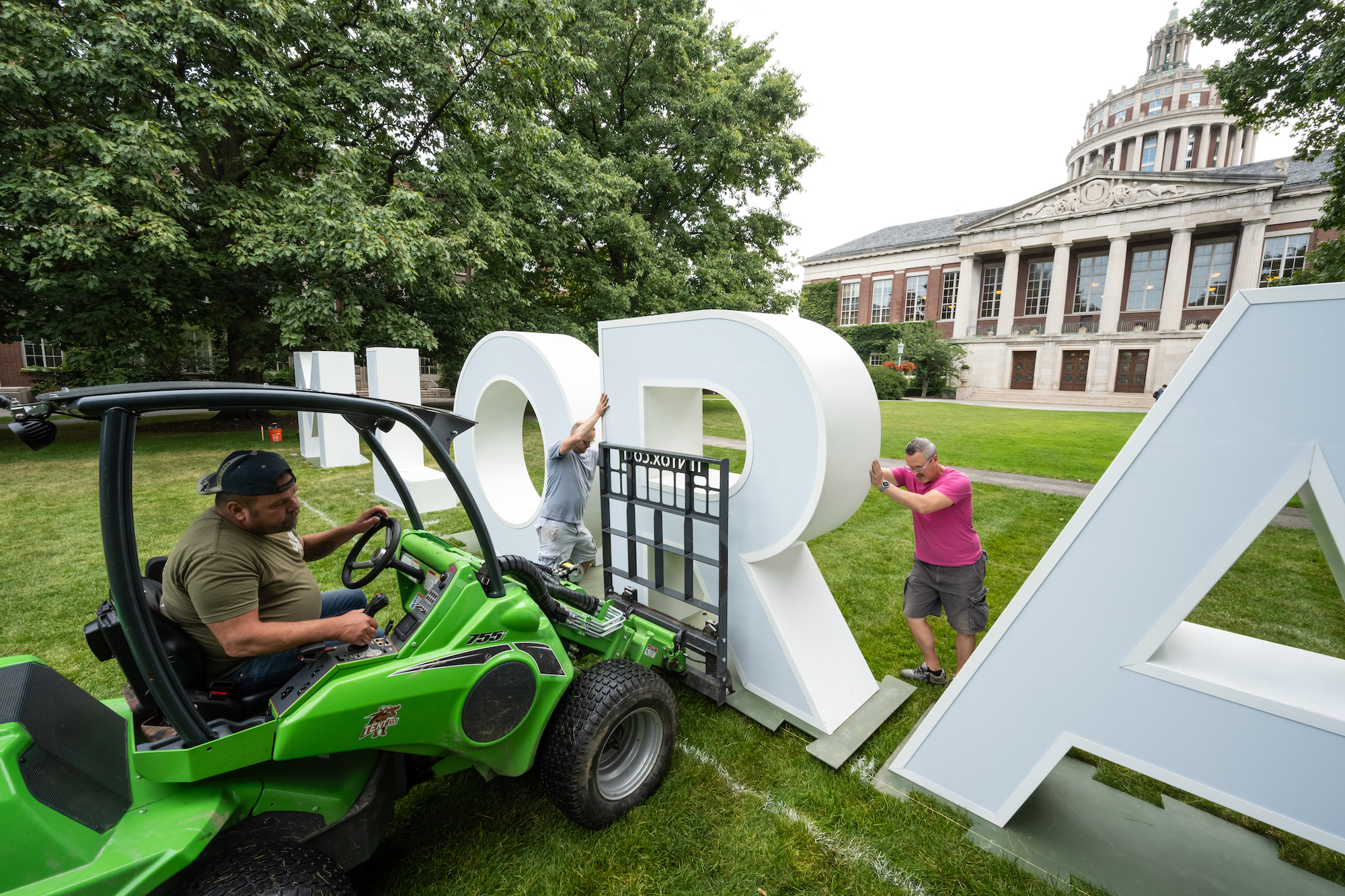 Large letters spelling MELIORA are positioned on the quad by three workers