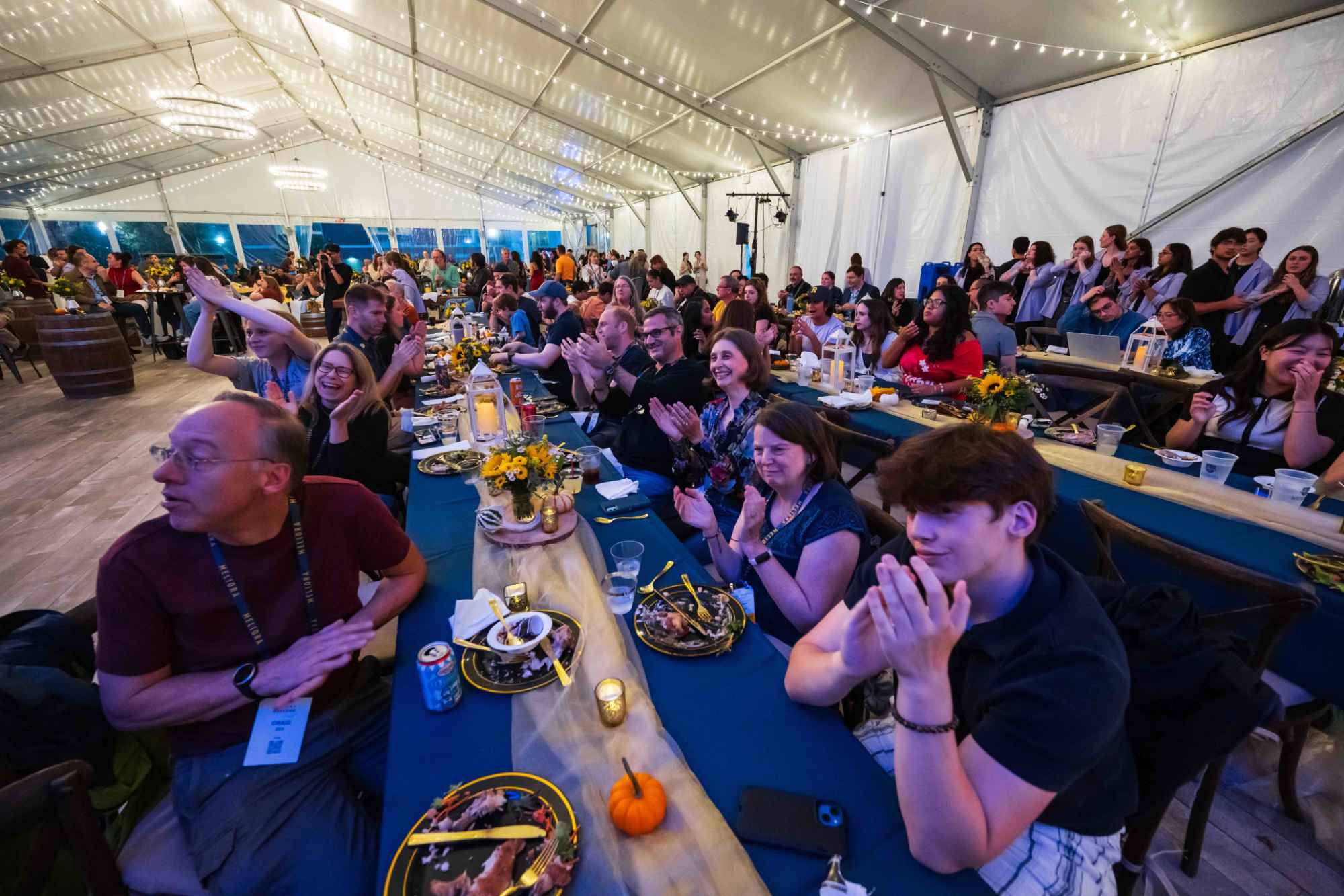 People seated at long rows of tables under the lights of a tent.
