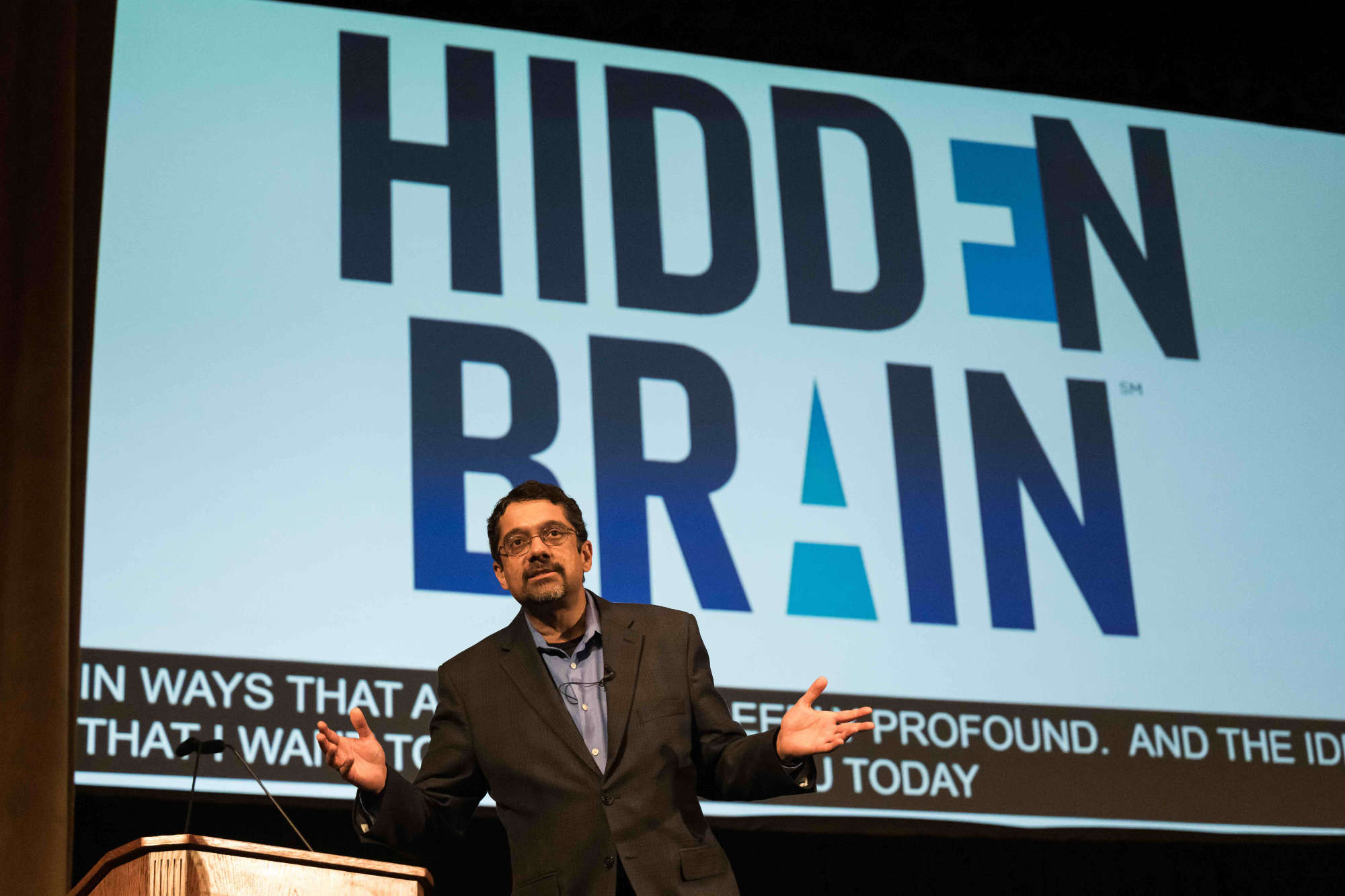 Shankar Vedantam presents on stage in front of a screen that says "Hidden Brain."