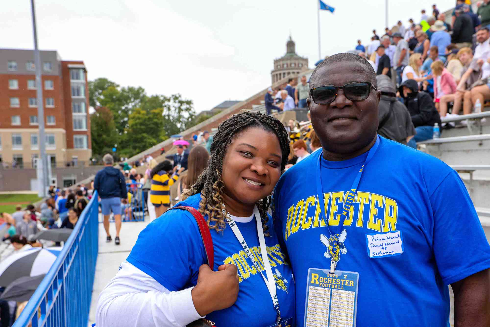 A mother and father in matching University of Rochester t-shirts smile for a photo with bleachers and campus in the background.