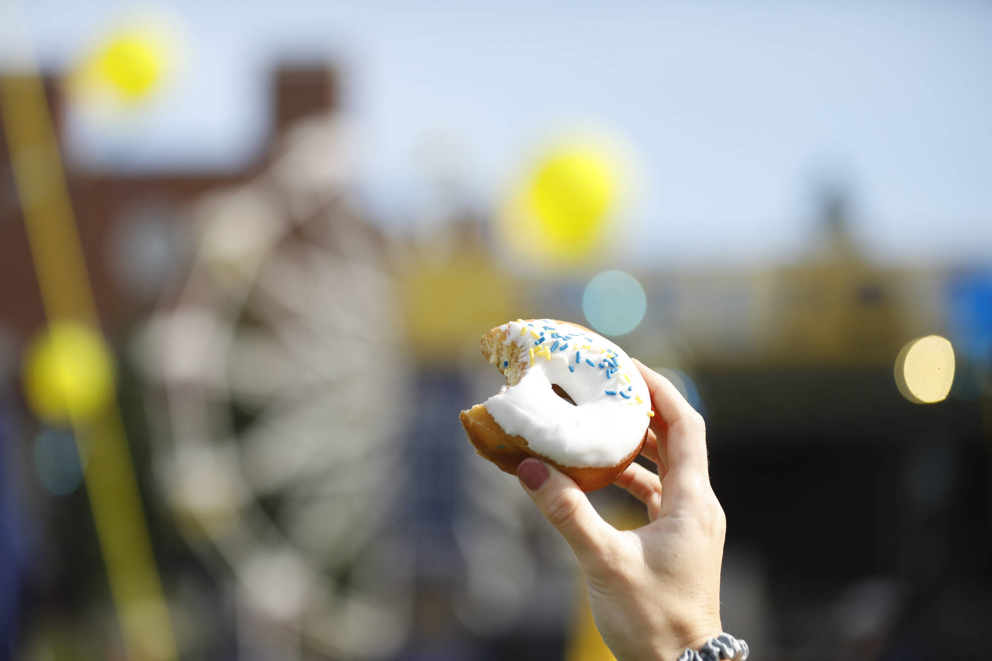 Hand holding a donut with white glaze and blue and yellow sprinkles with a ferris wheel blurred out in the background.