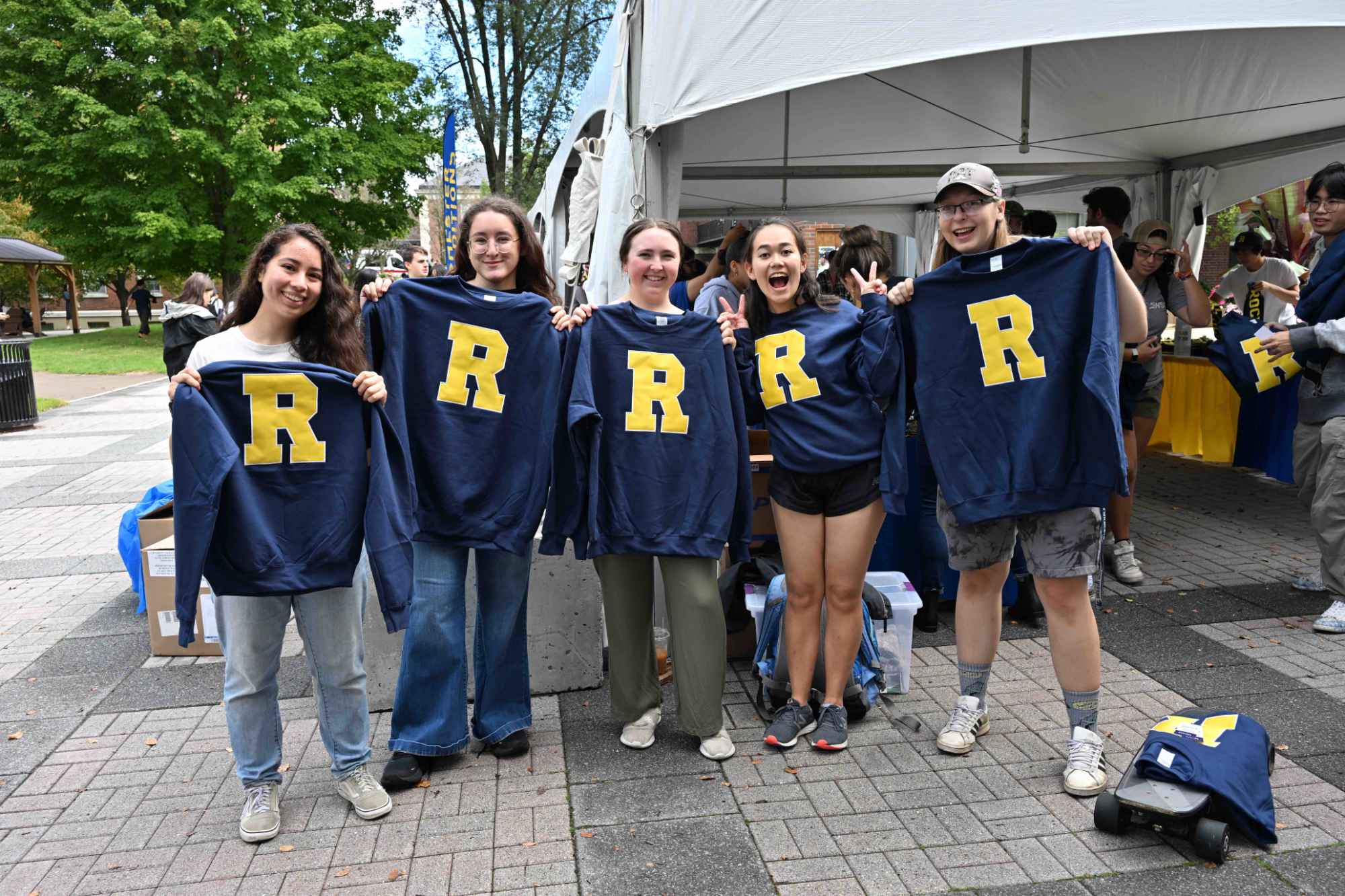 Five University of Rochester students smile and hold up blue sweatshirts with big yellow R's on them.