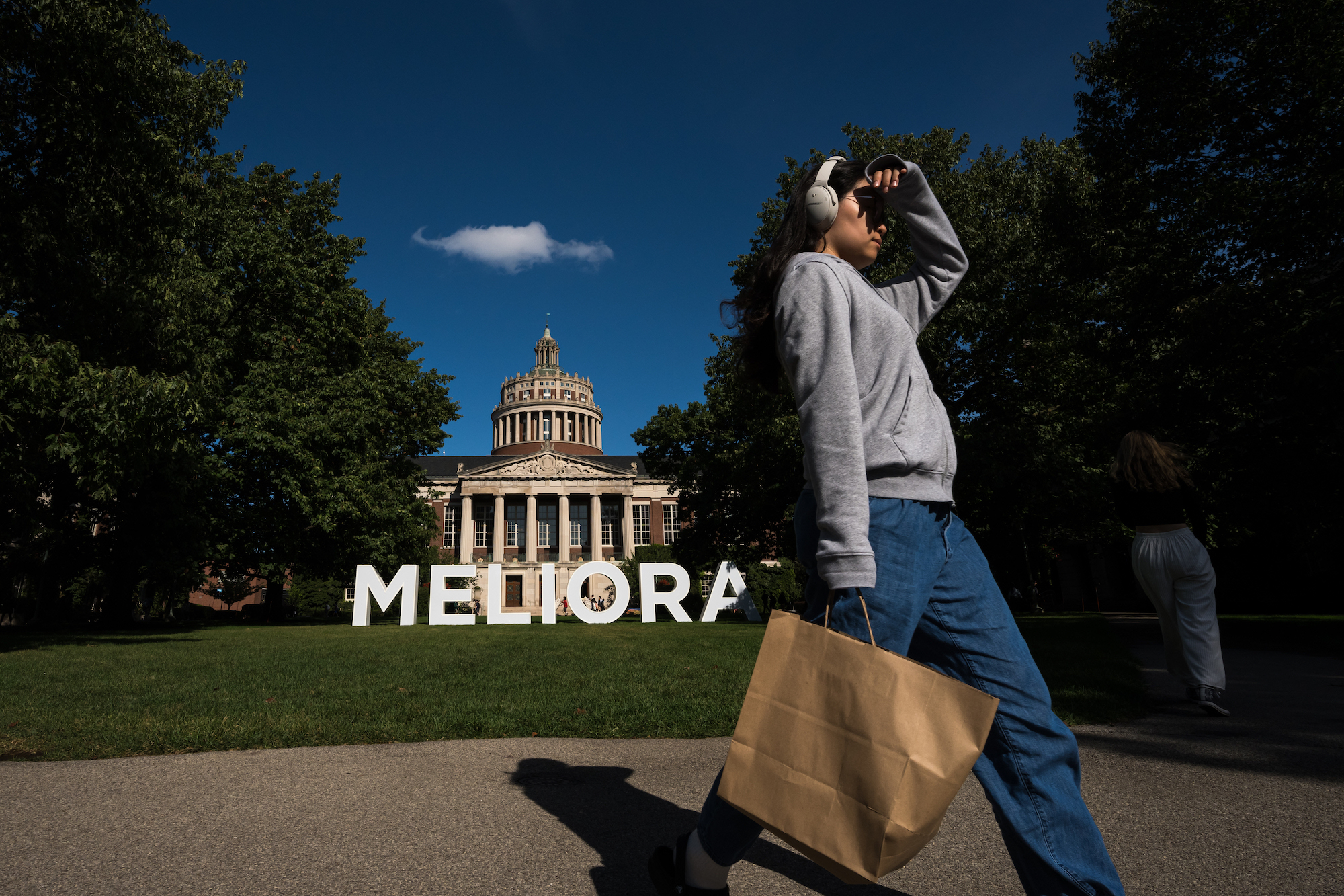 Student wearing headphones and carrying a paper shopping bag with large letters spelling MELIORA in the background