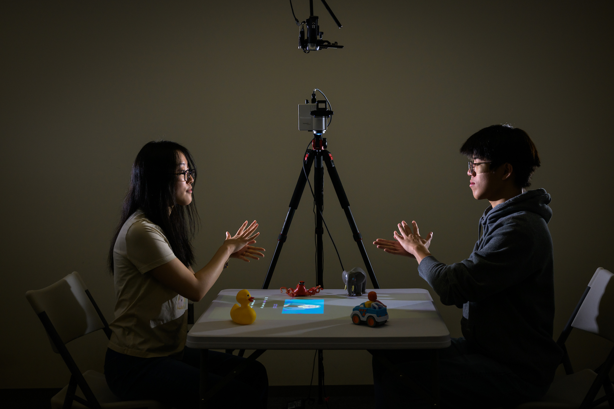 Two students sit across the table from one another. A device which uses augmented reality and artificial intelligence to help parents learn American Sign Language as they interact with their Deaf and hard-of-hearing children is between them