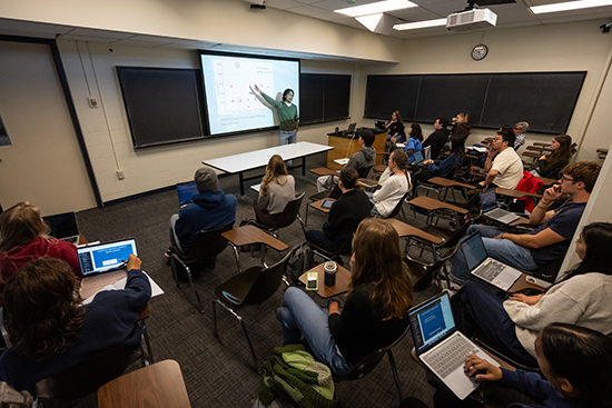 A professor stands in front of a classroom of students, pointing to a screen.