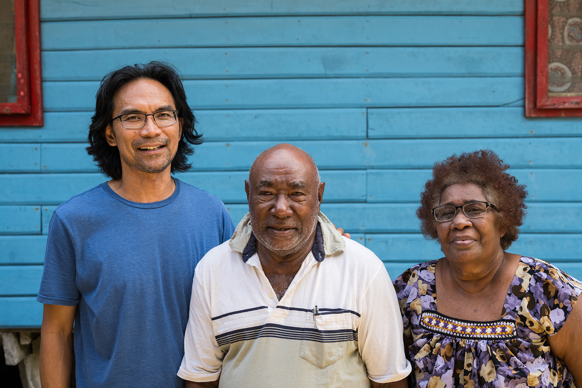 Three people, including a couple of elders, pose for a photo in front of a blue building.