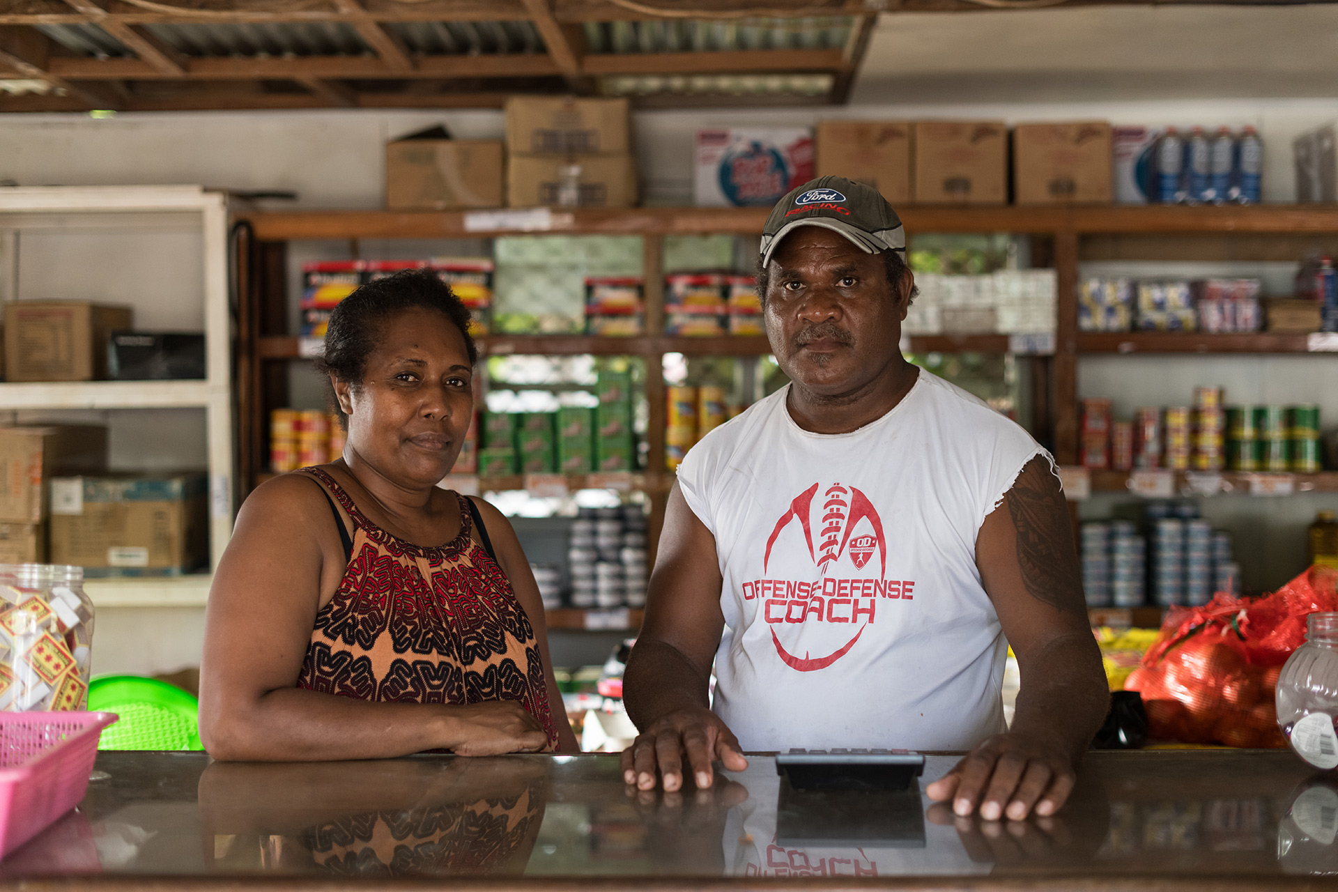 A couple stand behind the counter of a store, supplies stacked behind them.
