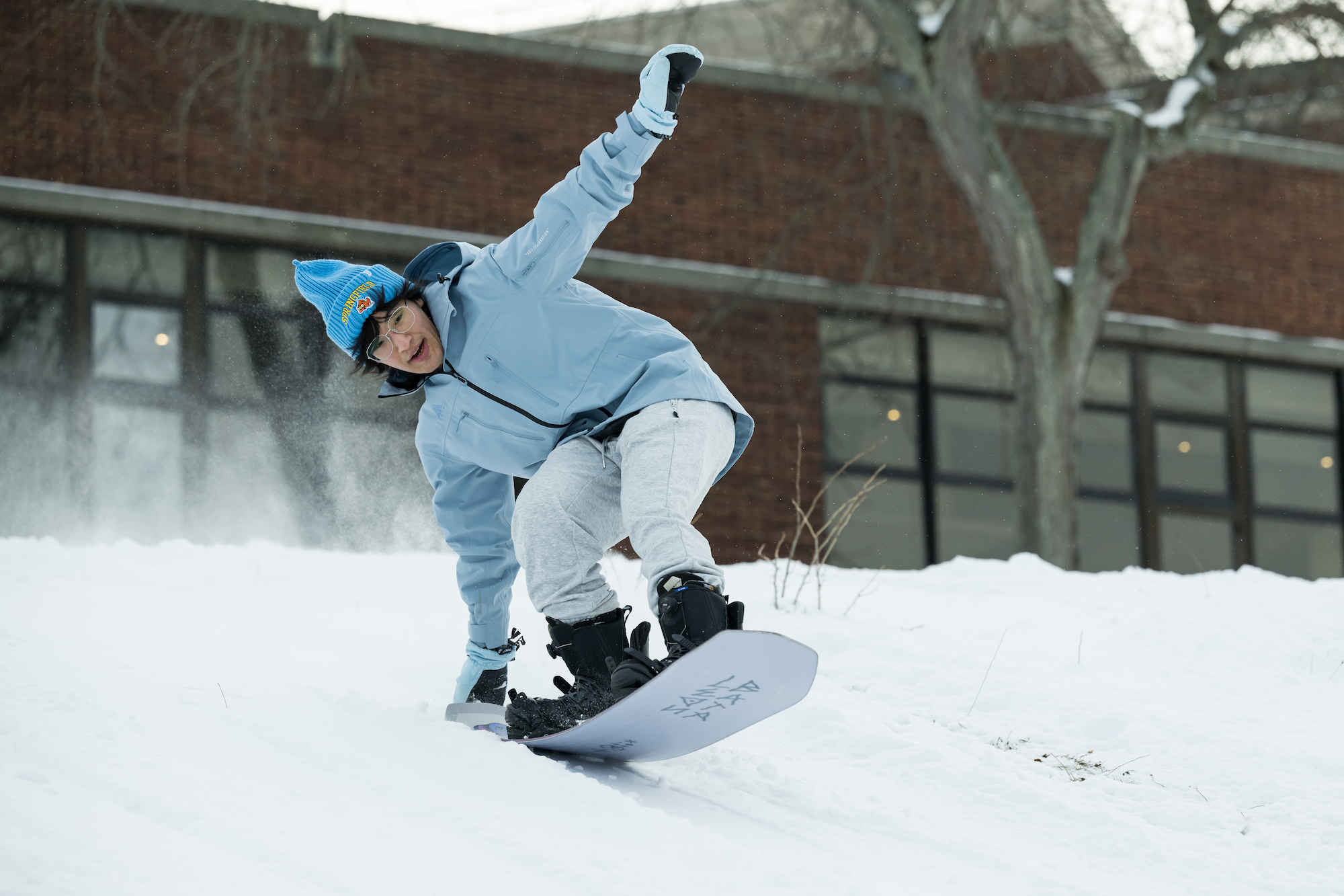 University of Rochester student in blue parka and white pants snowboards on the River Campus.