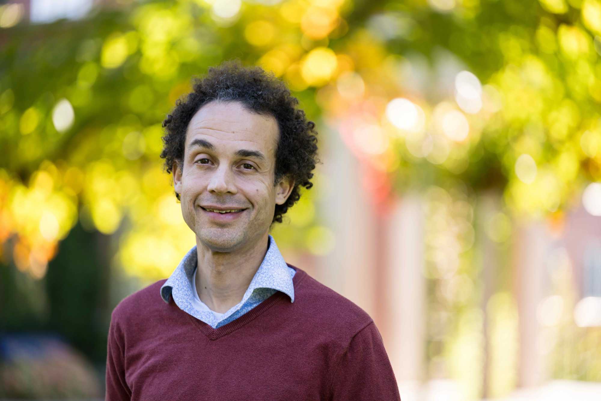 Mark Buckley in a collared shirt and red sweater smiles at the camera on the Eastman Quadrangle, surrounded by autumnal foliage.