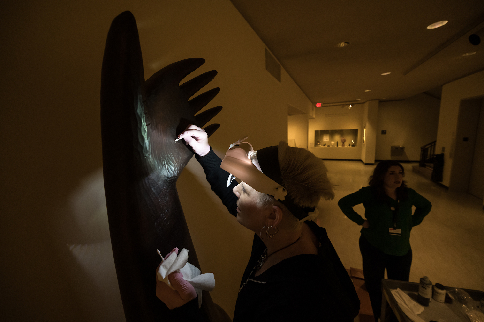 Person wearing headlamp cleans a large sculpture. Another employee stands nearby.