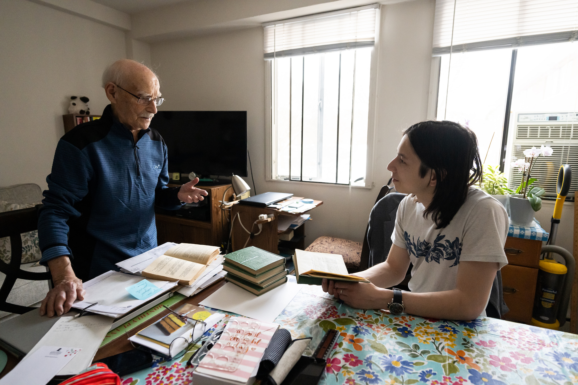 An older man speaks with a college student. The table in front of them is covered in books. 