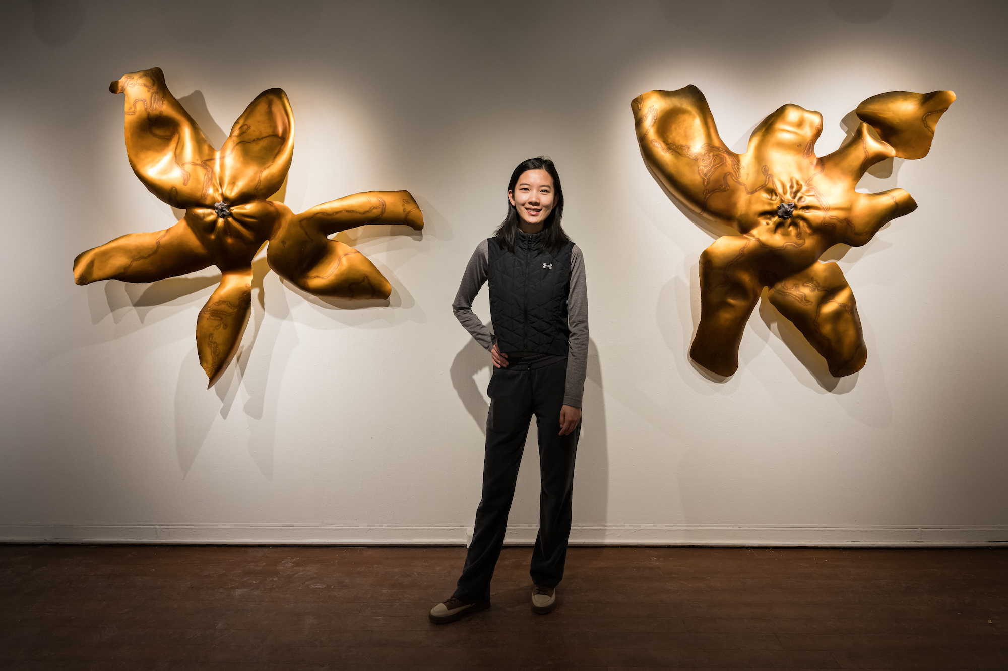 Artist is pictured in front of two large metallic sculptures hanging on a wall. 