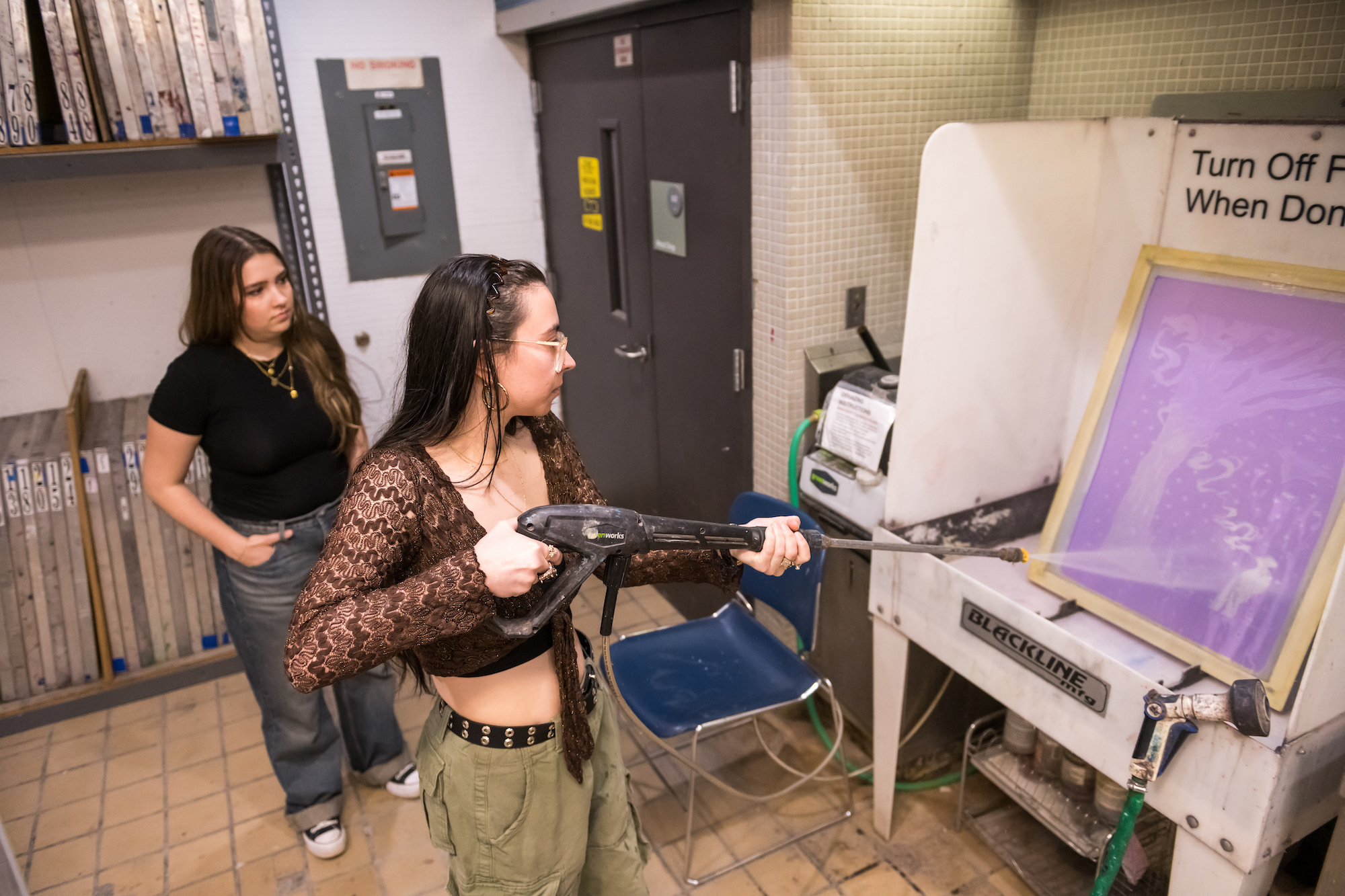 A student uses a power washer on a screen while another student looks on. 
