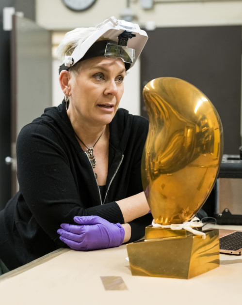 Kasia Maroney, wearing a headlamp, leans on a table with Jean Arp's Leaf on Crystal sculpture in the foreground.