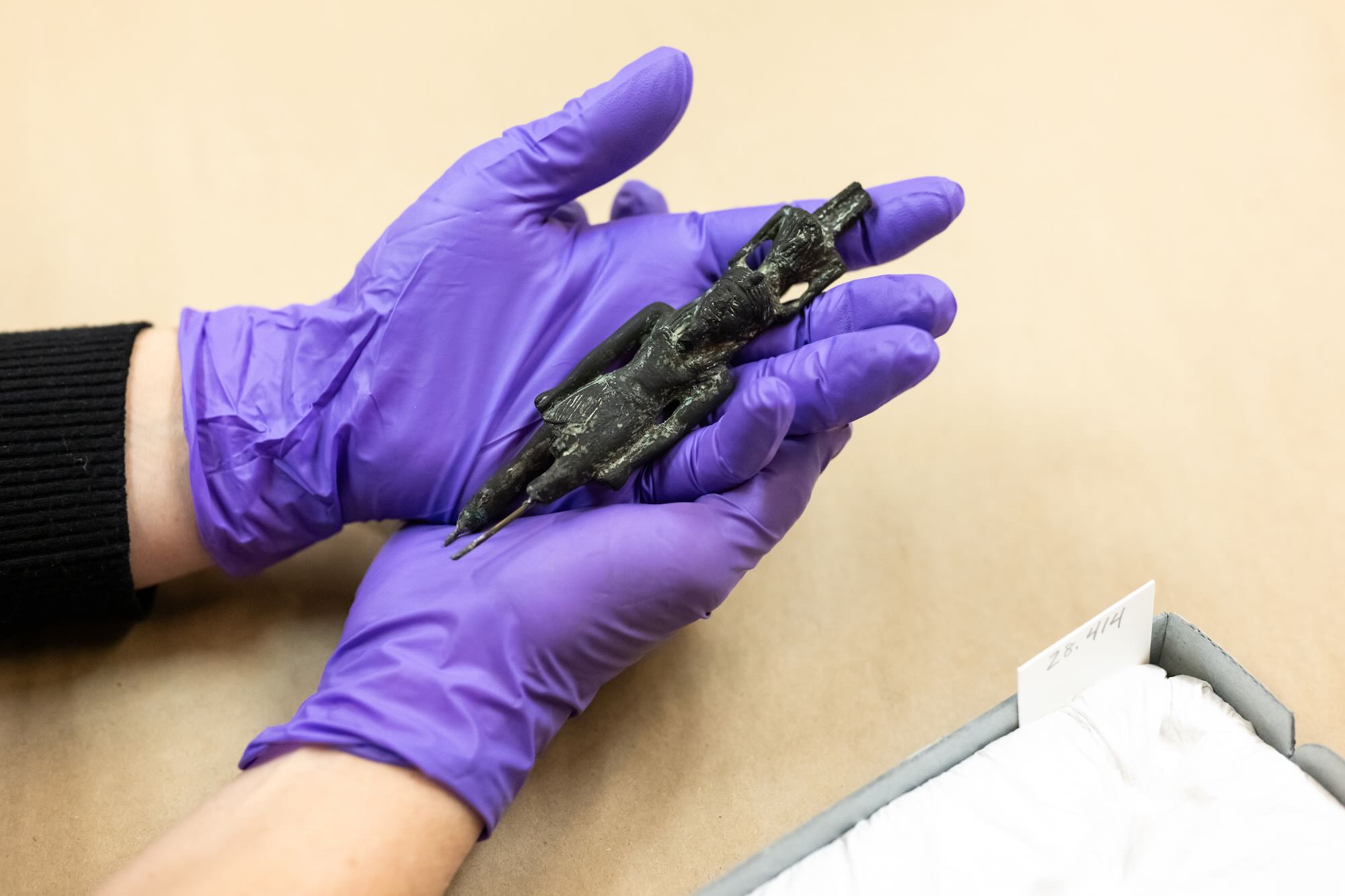 Two hands in purple latex gloves cradle a small oxidized bronze figure of Nefertem dating to ancient Egypt.