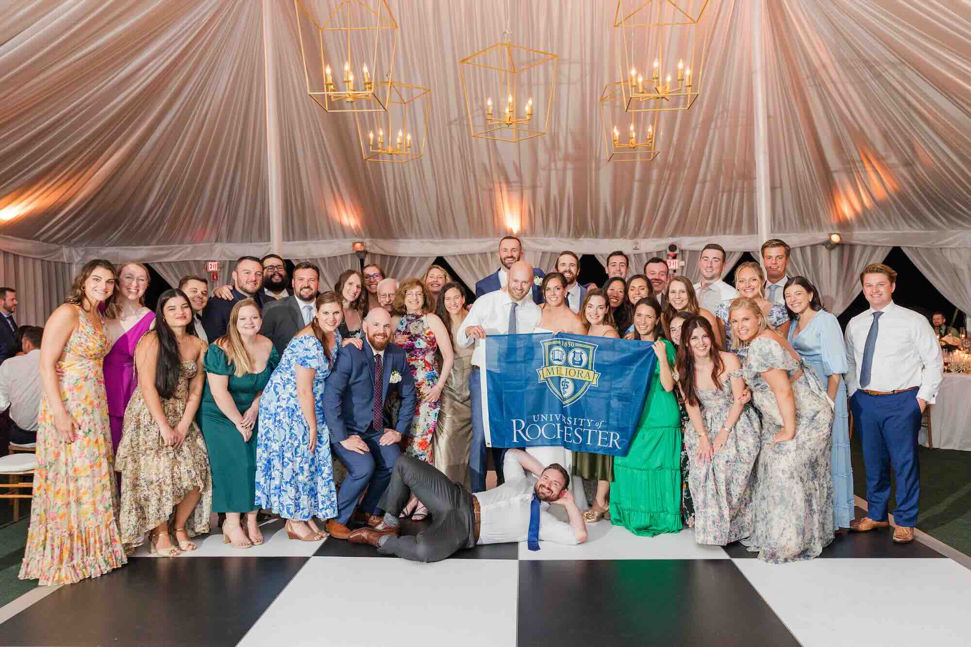 Large group photo at the wedding of Farrell Cooke and Tyler Viterise, with the couple at the center holding a University of Rochester flag