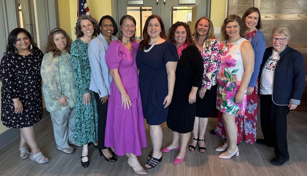 Twelve women dressed in bright, professional attire pose together indoors.