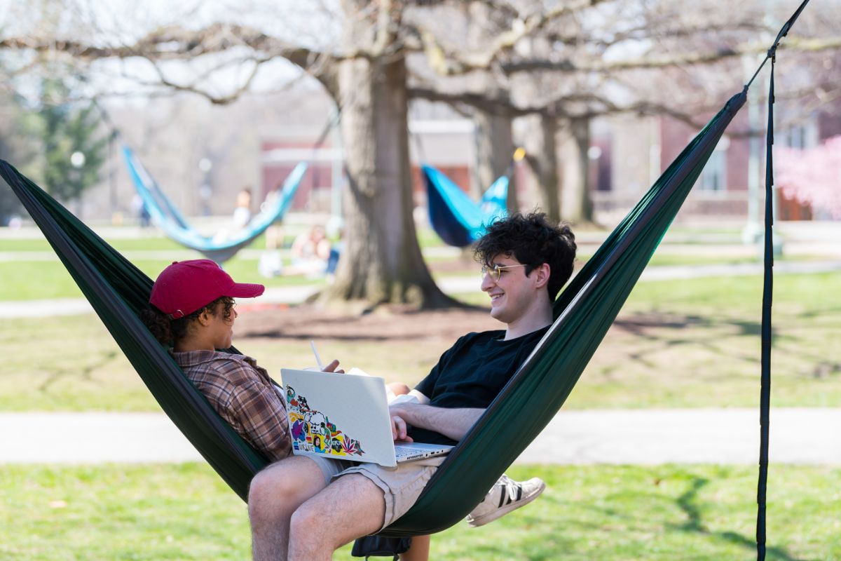 wo University of Rochester students relax in a hammock, working on laptops amidst a serene outdoor setting.
