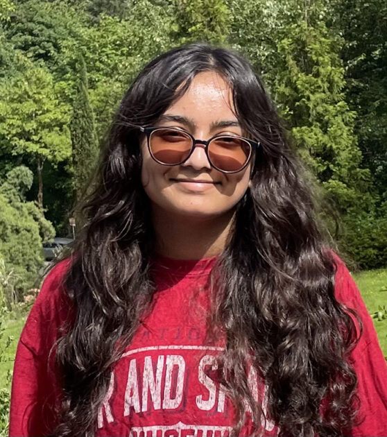 Student in sunglasses and a red T-shirt smiles outdoors on a sunny day, surrounded by lush green tree.