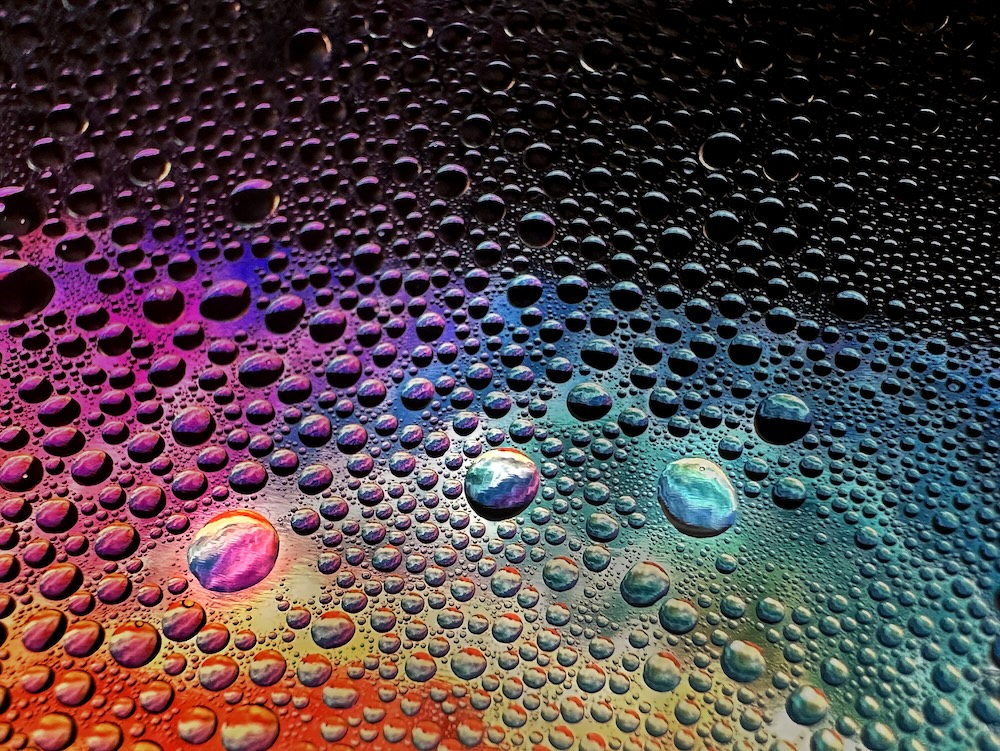 Close-up of multicolored water droplets on a surface, creating a galaxy-like pattern of vivid reds, purples, blues, and greens.