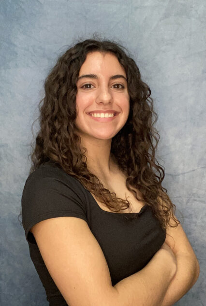Person with long curly hair smiling and posing with arms crossed in front of a blue backdrop.