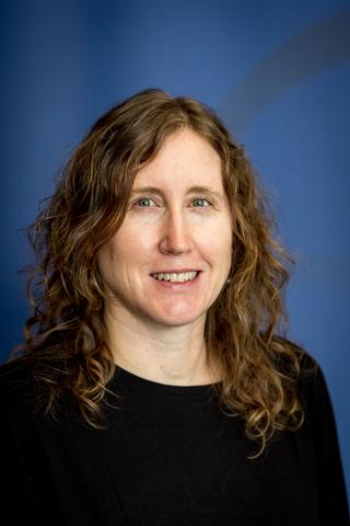 Portrait of a University of Rochester faculty member with wavy hair, wearing a black top, smiling in front of a blue background.