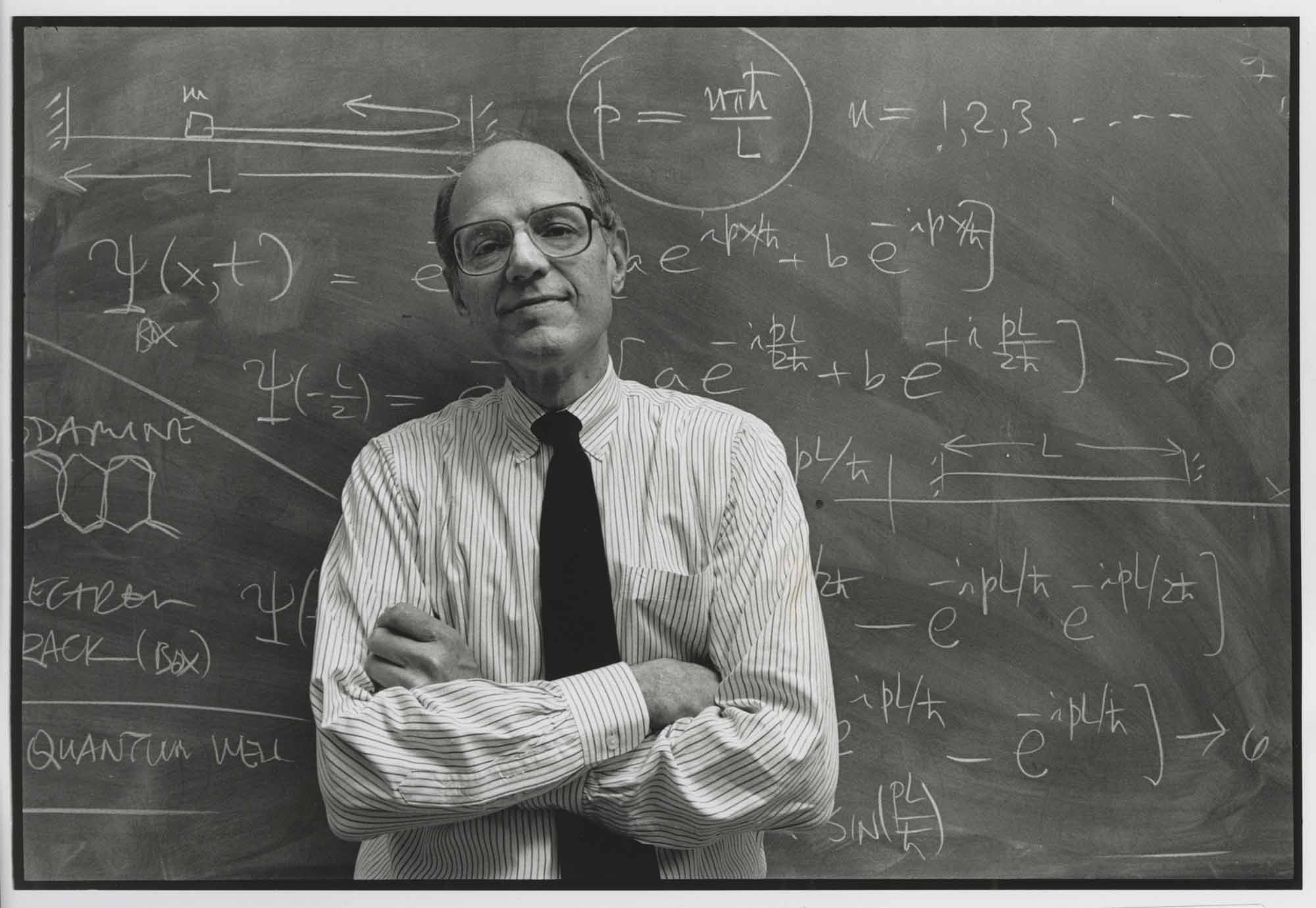 Black and white archival photo of Joseph Eberly in a shirt and tie leaning against a chalkboard covered in equations.