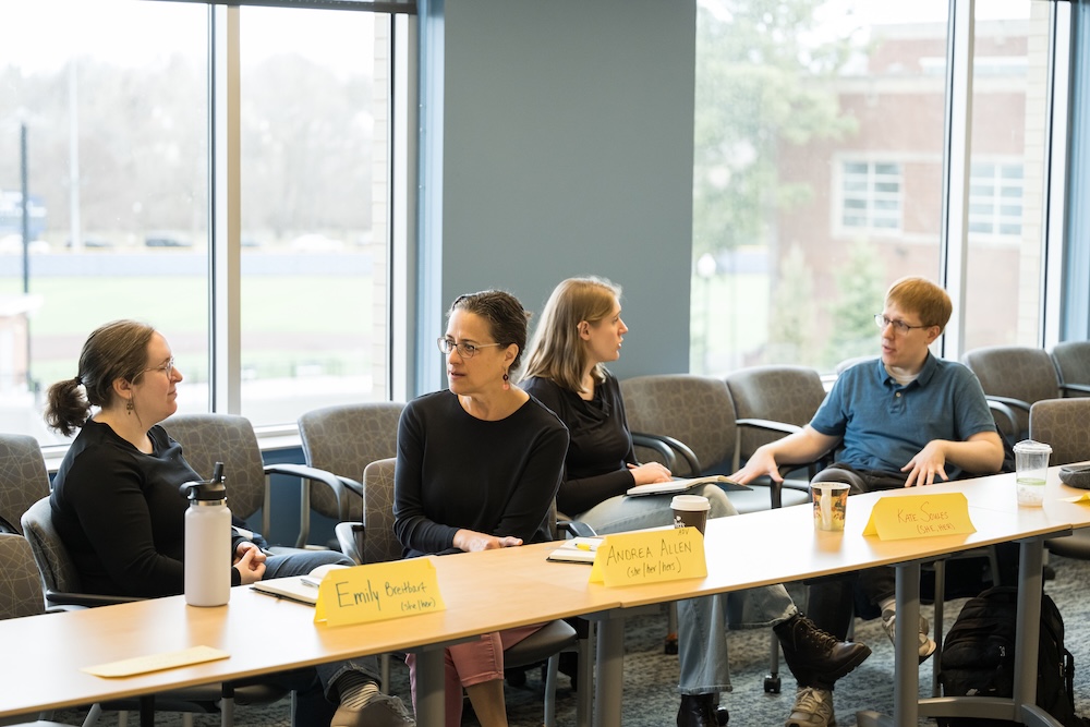 Four University of Rochester faculty members engage in discussion around a conference table with name cards and coffee cups, seated by large windows.