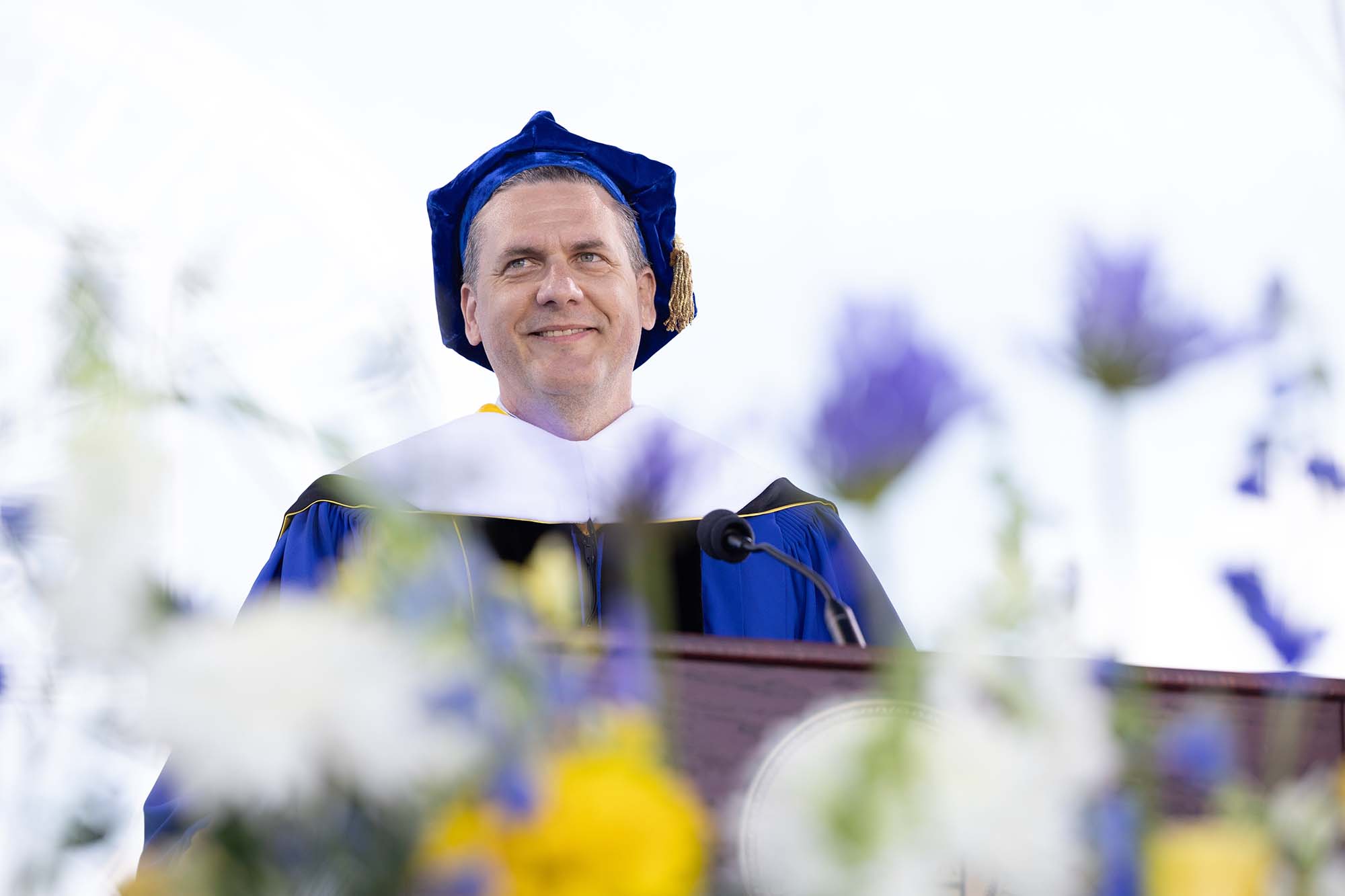 Tommy Evans in commencement regalia stands and smiles at a podium with flowers in the foreground. 