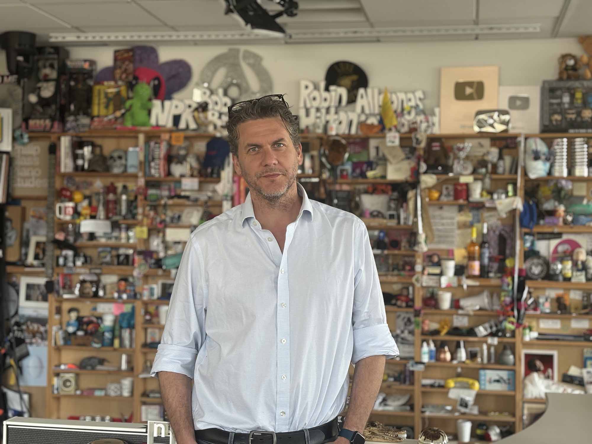 Tommy Evans in a button down shirt with the sleeves rolled up and his hands in his pocket looks at the camera from NPR's Tiny Desk studio. 