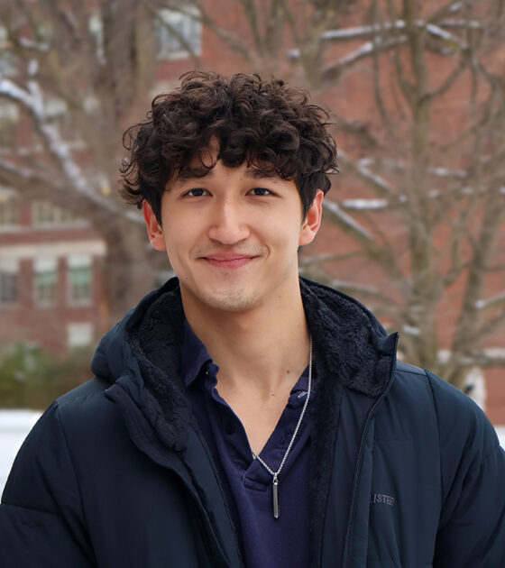 Smiling student in a winter coat stands outside on a snowy day with brick campus buildings and bare trees in the background.