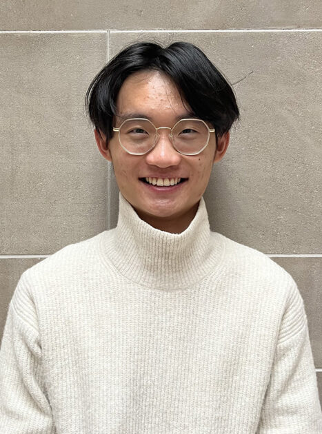 Person wearing glasses and a cream-colored turtleneck sweater smiling in front of a tiled wall.